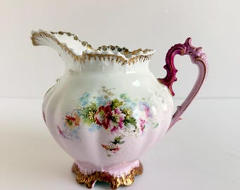 May include: A porcelain creamer featuring a white and soft pink gradient body with a hand-painted floral motif in shades of red, green, and blue. The piece has a scalloped rim with gold trim, a decorative magenta handle, and a gold-painted base.