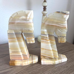May include: A pair of horse head bookends carved from layered beige and brown stone. The bookends are positioned on a wooden surface, with a lamp in the background.