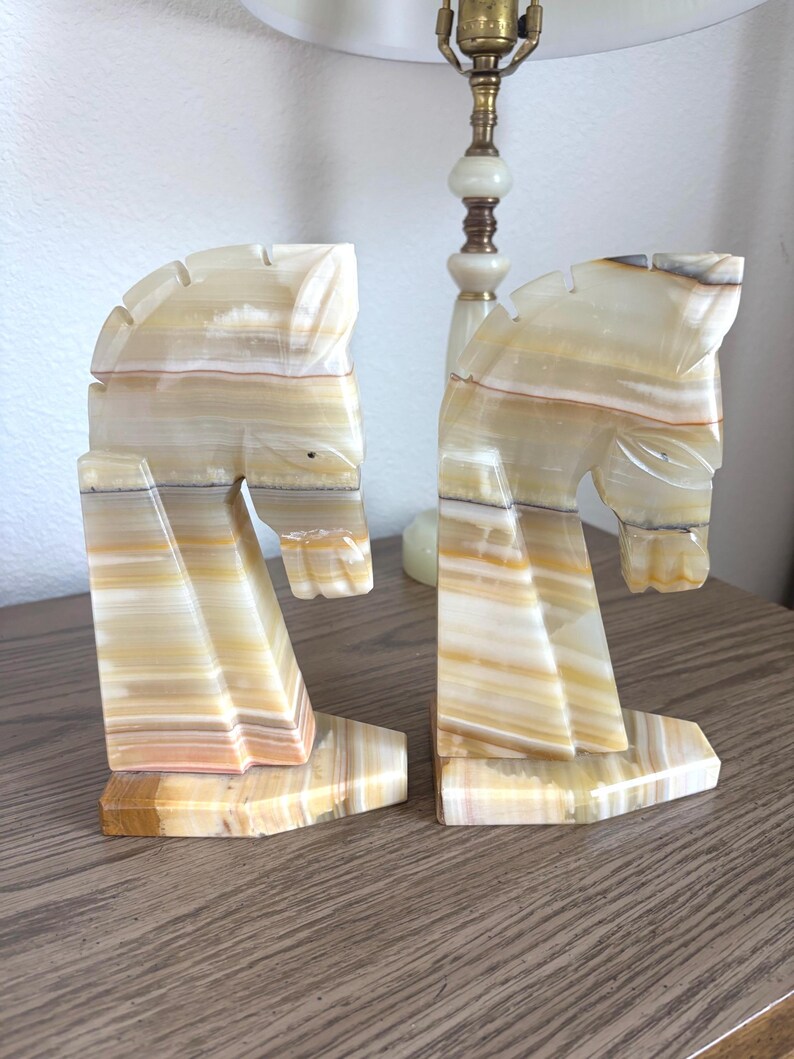 May include: A pair of horse head bookends carved from layered beige and brown stone. The bookends are positioned on a wooden surface, with a lamp in the background.