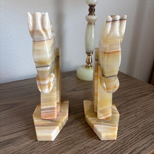 May include: A pair of onyx horse head bookends. The bookends are carved from stone with a cream, tan, and brown color scheme. The horse heads are detailed with a stylized mane and are mounted on a rectangular base.
