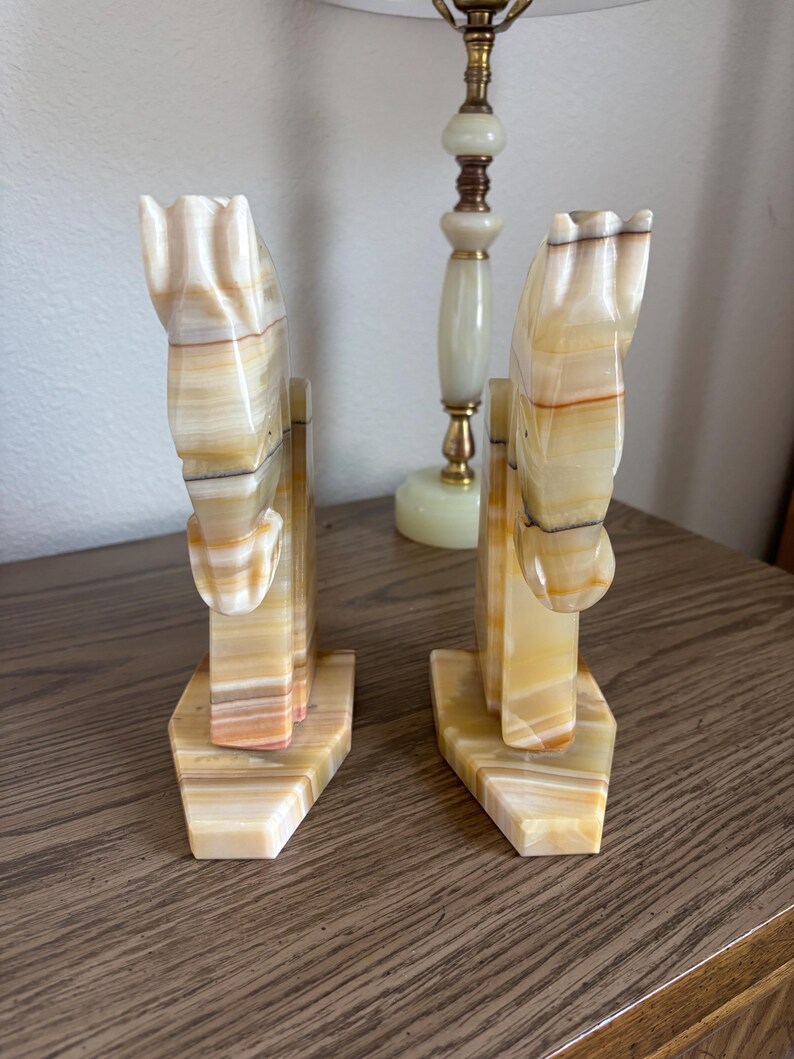 May include: A pair of onyx horse head bookends. The bookends are carved from stone with a cream, tan, and brown color scheme. The horse heads are detailed with a stylized mane and are mounted on a rectangular base.