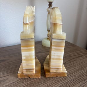May include: A pair of onyx bookends with a horse head design. The bookends are a mix of beige, brown, and white, with a rectangular base. The horse head is carved in a stylized manner. The bookends are a decorative item.
