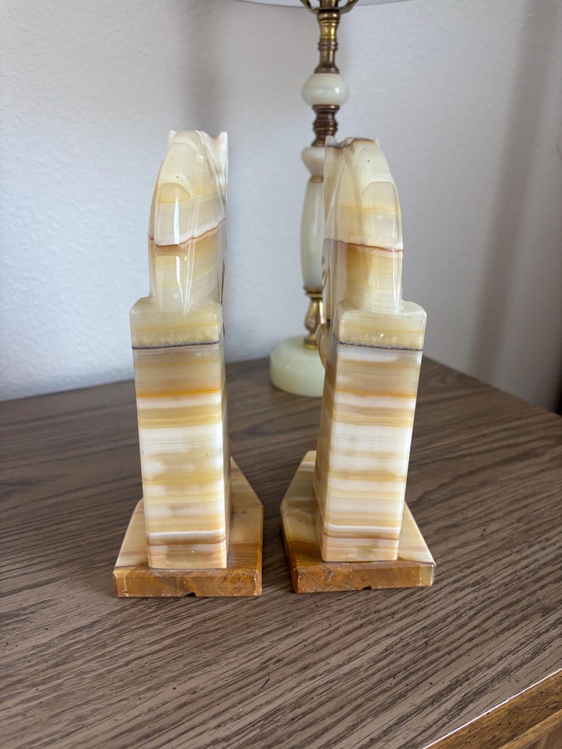 May include: A pair of onyx bookends with a horse head design. The bookends are a mix of beige, brown, and white, with a rectangular base. The horse head is carved in a stylized manner. The bookends are a decorative item.