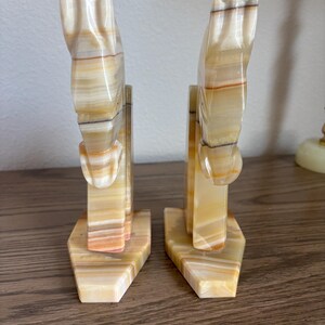 May include: A pair of decorative bookends carved from onyx. The bookends are shaped like horses, with a cream, tan, and brown striped pattern. The bases are hexagonal. The bookends are displayed on a wooden surface.