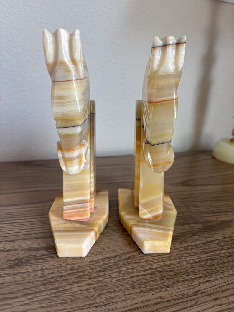 May include: A pair of decorative bookends carved from onyx. The bookends are shaped like horses, with a cream, tan, and brown striped pattern. The bases are hexagonal. The bookends are displayed on a wooden surface.