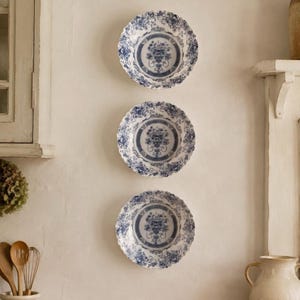May include: Three round ceramic plates arranged vertically, each featuring a scalloped edge and a blue and white transferware pattern. The design includes a central floral urn motif surrounded by intricate floral borders and lattice detailing.