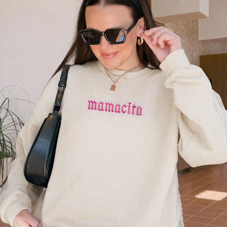 May include: Cream-colored sweatshirt with the embroidered word "mamacita" in pink. The sweatshirt has long sleeves and a crew neck. A black shoulder bag is visible on the left side of the image. The person in the image is wearing black sunglasses and gold jewelry.