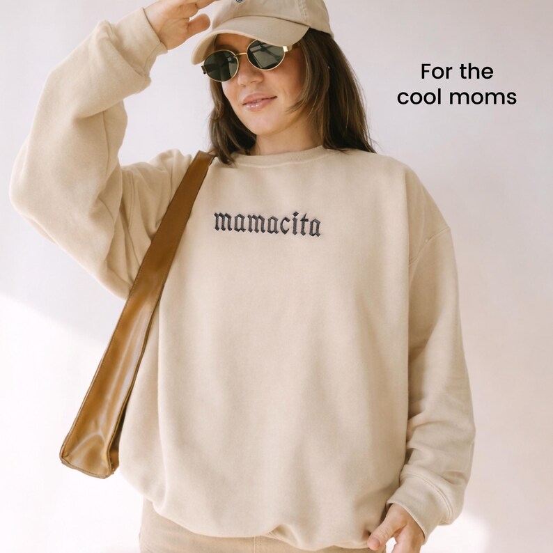 May include: Beige sweatshirt with the word "mamacita" embroidered in black script. The image also includes a beige baseball cap, sunglasses, and a brown shoulder bag. Text on the image reads "For the cool moms."