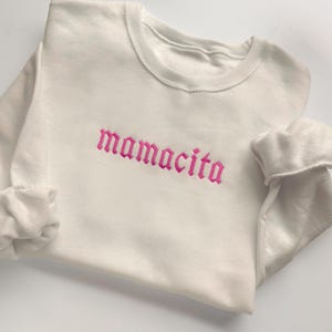 May include: Cream-colored sweatshirt with the word "mamacita" embroidered in pink gothic lettering across the chest. The sweatshirt has a crew neck and long sleeves with folded cuffs. The fabric appears soft and comfortable.