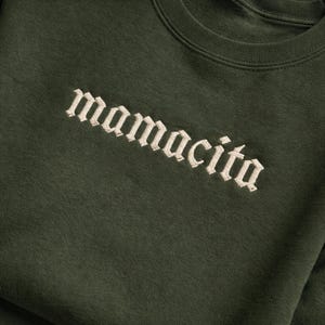 May include: Dark green sweatshirt with the word "mamacita" embroidered in a cream-colored gothic font. The text is diagonally aligned across the chest of the garment. The sweatshirt has a crew neck.