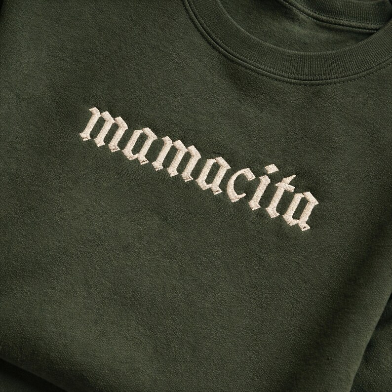 May include: Dark green sweatshirt with the word "mamacita" embroidered in a cream-colored gothic font. The text is diagonally aligned across the chest of the garment. The sweatshirt has a crew neck.