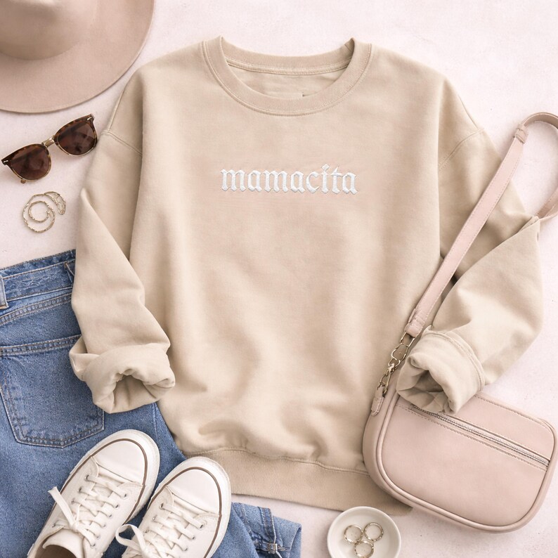 May include: Beige sweatshirt with the word "mamacita" embroidered in white. The sweatshirt is paired with blue jeans, white sneakers, a pink purse, sunglasses, and a hat. A casual, everyday outfit.