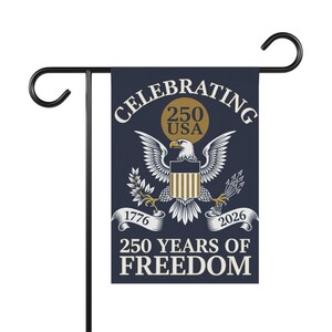 Puede incluir: Una bandera de jardín azul marino sobre un soporte de metal negro. La bandera presenta un círculo dorado con "250 USA" y un emblema de águila blanca. El texto dice "CELEBRATING 250 YEARS OF FREEDOM" con "1776" y "2026" en la parte inferior.