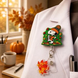 May include: A badge reel featuring a glittery green background with a cartoon dog wearing an orange hat and scarf, surrounded by autumn leaves. A clear plastic strap connects to a separate acrylic charm shaped like a vibrant orange and red maple leaf.