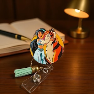 May include: A circular badge reel featuring a colorful illustration of a woman with long black hair and a large orange and black tiger. The badge reel includes a light mint green fabric tassel attached by a gold-toned metal cap and a clear strap.