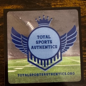 May include: A square sticker with a blue and white logo for Total Sports Authentics. The logo features a crown, wings, and the text "TOTAL SPORTS AUTHENTICS" in a circular design. The background shows a football field.
