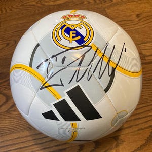 Puede incluir: Un bal&oacute;n de f&uacute;tbol blanco con el escudo del club Real Madrid en azul, dorado y blanco, un logotipo de Adidas negro y l&iacute;neas amarillas. El bal&oacute;n incluye una firma destacada en tinta negra en el centro y el texto Match Ball Replica Club.