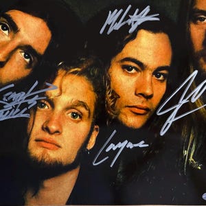 May include: A color photograph featuring four individuals with long hair, arranged in a close-up composition. The image includes handwritten signatures in silver ink across the faces and a small authentication sticker in the bottom right corner.