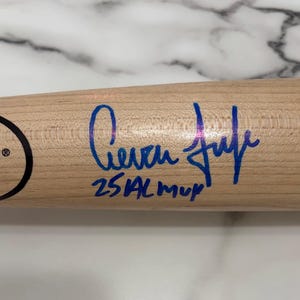 May include: A light-colored wooden baseball bat featuring a blue ink autograph and the handwritten text 25 AL MVP. A partial black logo is visible on the left side of the bat, displaying the word LE and the location LE, KY in small print.