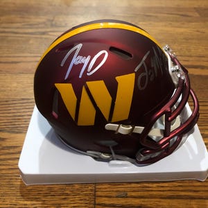 May include: A burgundy and gold American football helmet with the letter "W" in gold. The helmet has a gold stripe and a burgundy face mask. The helmet is on a white stand and has two signatures in white.