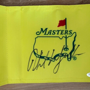 May include: A bright yellow golf flag featuring the green Masters logo with a map of the United States and a red flag pin. The fabric displays a prominent black ink autograph across the centre and a small white authentication sticker in the corner.