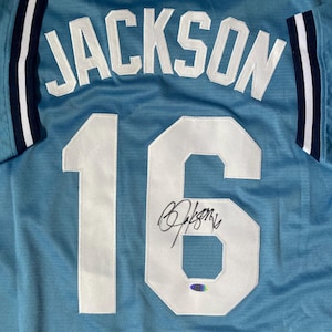 May include: Light blue jersey with the name "JACKSON" in white block letters across the back. The number "16" is also in white, with a signature above a holographic sticker. The sleeves have white and navy blue stripes.