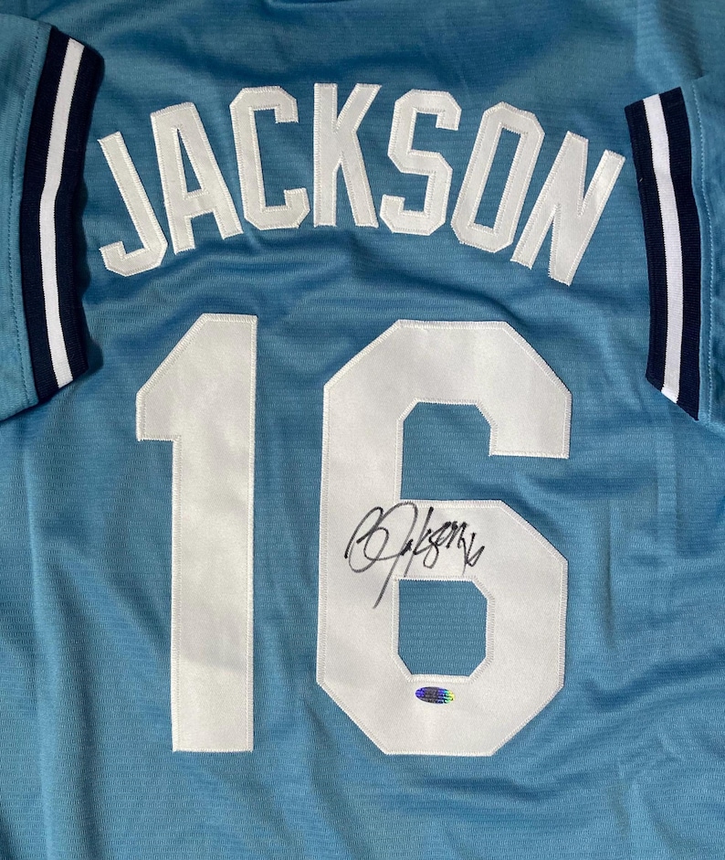May include: Light blue jersey with the name "JACKSON" in white block letters across the back. The number "16" is also in white, with a signature above a holographic sticker. The sleeves have white and navy blue stripes.