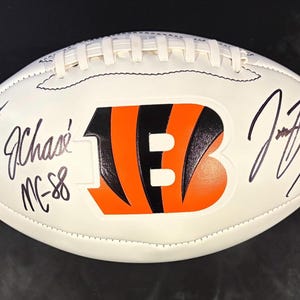 May include: A white football featuring the orange and black Cincinnati Bengals logo in the center. The surface is covered in multiple black ink signatures and the text "JC-88". The ball has white laces along the top and a textured leather exterior.