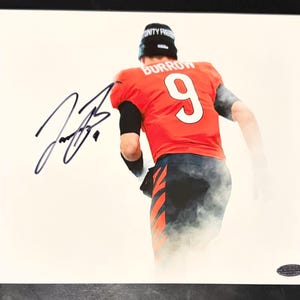 Op de afbeelding: Een foto van een footballspeler in een oranje shirt met de naam BURROW en het nummer 9 op de rug, gecombineerd met een zwarte muts. De afbeelding bevat een opvallende handtekening in zwarte inkt aan de linkerkant en een kleine authenticatiesticker.