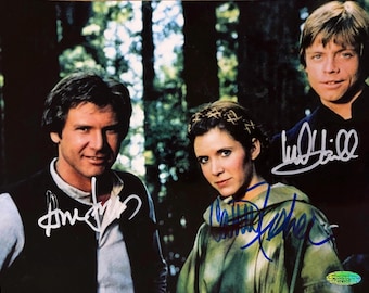 May include: A color photograph featuring three individuals from a film, with Harrison Ford on the left, Carrie Fisher in the center, and Mark Hamill on the right. Each person has a signature written in ink across their image on the print.