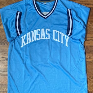 May include: Light blue jersey with the words "KANSAS CITY" in white across the front. The jersey has a V-neck with white and navy blue trim, and navy blue and white stripes on the sleeves.