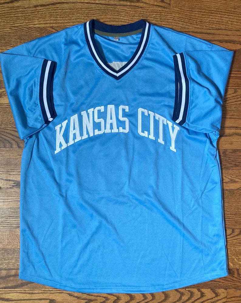 May include: Light blue jersey with the words "KANSAS CITY" in white across the front. The jersey has a V-neck with white and navy blue trim, and navy blue and white stripes on the sleeves.