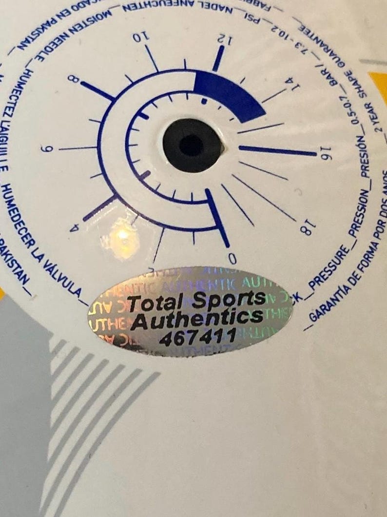 Puede incluir: Una superficie circular blanca con un gr&aacute;fico de man&oacute;metro azul con marcas de 0 a 18 PSI. Una pegatina hologr&aacute;fica ovalada est&aacute; centrada en la superficie, mostrando el texto Total Sports Authentics y el n&uacute;mero 467411 en negro.