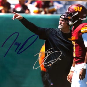 May include: An autographed photo featuring a football coach pointing and a player in a burgundy and gold uniform. The player wears a helmet with a gold "W" logo. The photo has two signatures in blue and silver ink.