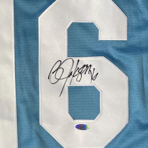 May include: Light blue jersey with a large white number 6 patch. The number is stitched onto the jersey and has a signature in black ink across the number. A holographic sticker is below the signature.