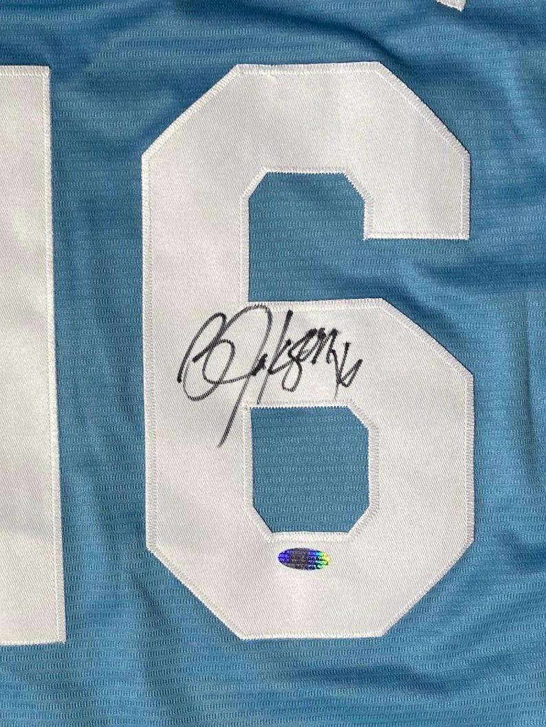 May include: Light blue jersey with a large white number 6 patch. The number is stitched onto the jersey and has a signature in black ink across the number. A holographic sticker is below the signature.