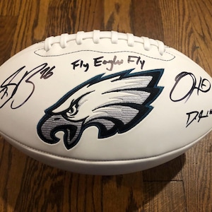 Puede incluir: Balón de fútbol blanco con el logotipo de los Eagles de Filadelfia y la frase "Fly Eagles Fly". El balón está firmado por varias personas. El balón está sobre una superficie de madera.