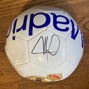 May include: A white soccer ball featuring blue text that reads madrid and a colorful team crest with a crown. The surface of the ball displays a black ink autograph signature centered between the panels, showcasing a classic multi-paneled design.