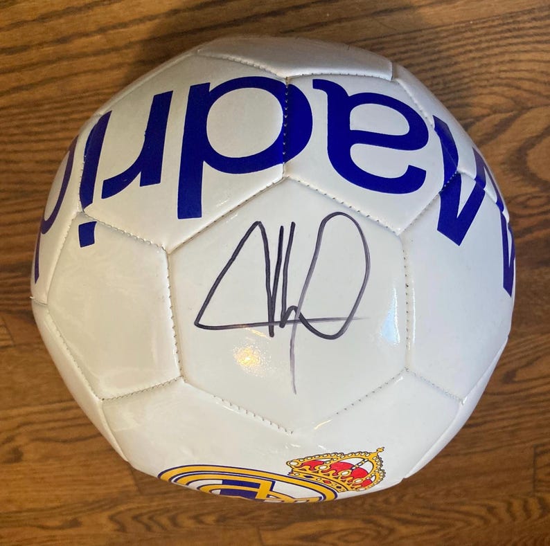 May include: A white soccer ball featuring blue text that reads madrid and a colorful team crest with a crown. The surface of the ball displays a black ink autograph signature centered between the panels, showcasing a classic multi-paneled design.