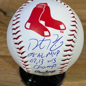 Può includere: Una palla da baseball bianca con cuciture rosse, caratterizzata da un logo rosso dei Boston Red Sox e una firma a inchiostro blu. Sotto la firma, il testo recita '08 AL MVP' e '07, 13 WS Champ'. Il marchio Rawlings è stampato in nero in basso.