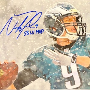 Può includere: Una fotografia di un giocatore di football americano con una maglia color verde acqua dei Philadelphia Eagles con il numero 9, che indossa un casco e tiene un pallone durante una nevicata. L'immagine presenta un autografo in inchiostro blu e la scritta SB LII MVP.