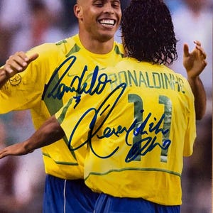 Puede incluir: Una fotografía de dos jugadores de fútbol con camisetas amarillas con detalles verdes y pantalones cortos azules. El jugador en primer plano tiene el nombre RONALDINHO y el número 11 en la espalda. Ambas camisetas tienen autógrafos en tinta azul.