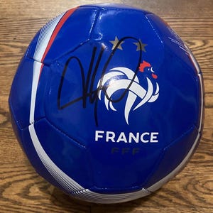 Può includere: Un pallone da calcio blu con il logo bianco France FFF e due stelle dorate sopra. Il pallone presenta accenti grafici bianchi e rossi e una firma a inchiostro nero sulla superficie. Il testo FRANCE è stampato in lettere maiuscole bianche.