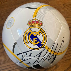 Puede incluir: Un bal&oacute;n de f&uacute;tbol blanco con el escudo del Real Madrid en amarillo, azul y rojo con una corona en la parte superior. El bal&oacute;n tiene detalles gr&aacute;ficos en gris y amarillo y una firma en tinta negra en la parte inferior de la superficie.