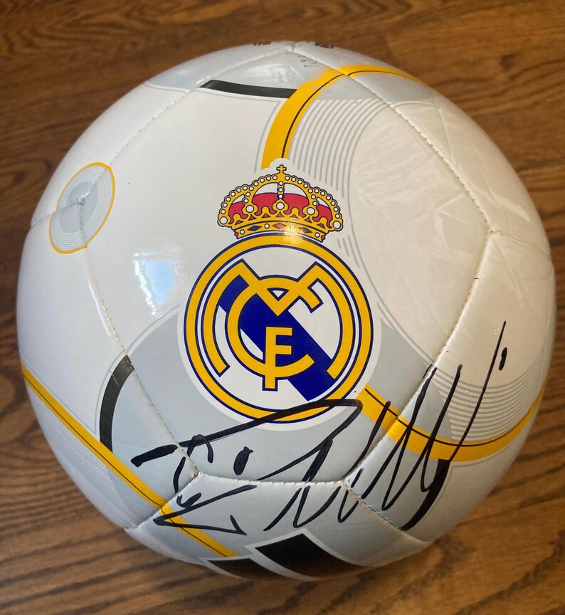Puede incluir: Un bal&oacute;n de f&uacute;tbol blanco con el escudo del Real Madrid en amarillo, azul y rojo con una corona en la parte superior. El bal&oacute;n tiene detalles gr&aacute;ficos en gris y amarillo y una firma en tinta negra en la parte inferior de la superficie.