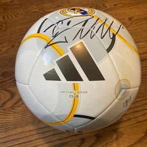 Puede incluir: Un bal&oacute;n de f&uacute;tbol Adidas blanco con detalles gr&aacute;ficos en gris y amarillo, el escudo del club Real Madrid y el texto MATCH BALL REPLICA CLUB. Una firma en tinta negra se muestra claramente en la parte superior de la superficie del bal&oacute;n.