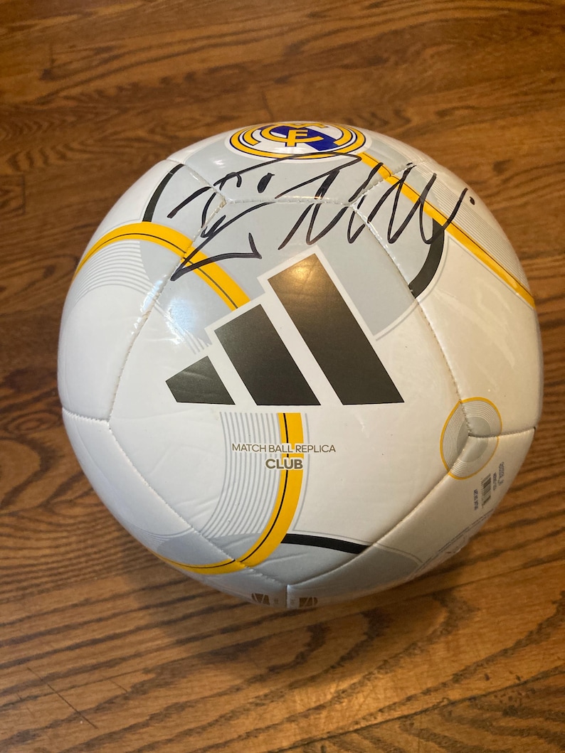 Puede incluir: Un bal&oacute;n de f&uacute;tbol Adidas blanco con detalles gr&aacute;ficos en gris y amarillo, el escudo del club Real Madrid y el texto MATCH BALL REPLICA CLUB. Una firma en tinta negra se muestra claramente en la parte superior de la superficie del bal&oacute;n.
