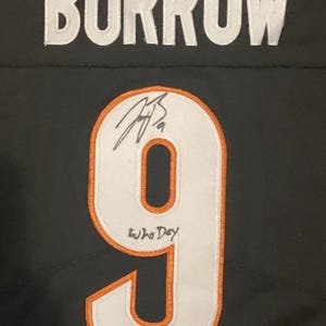 Op de afbeelding: Een zwart sportshirt met de naam BURROW in witte letters boven het nummer 9. Het nummer 9 is wit met een oranje rand en bevat een handtekening in zwarte inkt, de handgeschreven tekst Who Dey en een kleine zilverkleurige sticker.