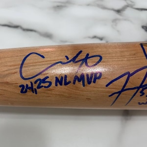 May include: A light-colored wooden baseball bat featuring blue ink signatures and handwritten inscriptions. One inscription clearly reads 24, 25 NL MVP, while another signature includes the text 20 WS, all written directly onto the smooth wood surface.