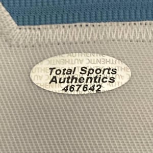 May include: Close-up of a light gray fabric with a white stitched edge and a blue fabric in the background. An oval sticker with the text "Total Sports Authentics" and the number "467642" is visible.